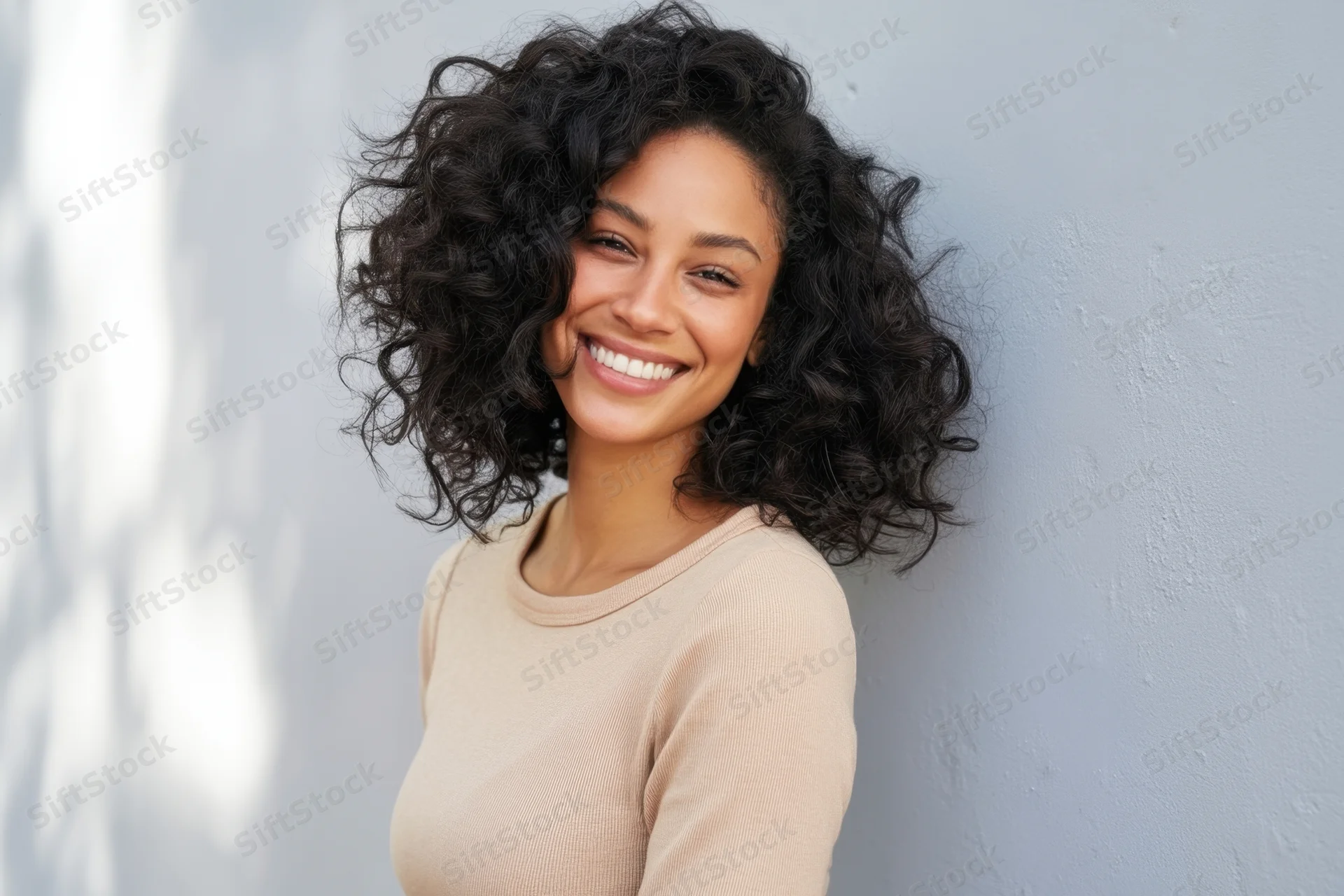 Smiling woman with curly black hair wearing beige sweater leaning against gray wall outdoors