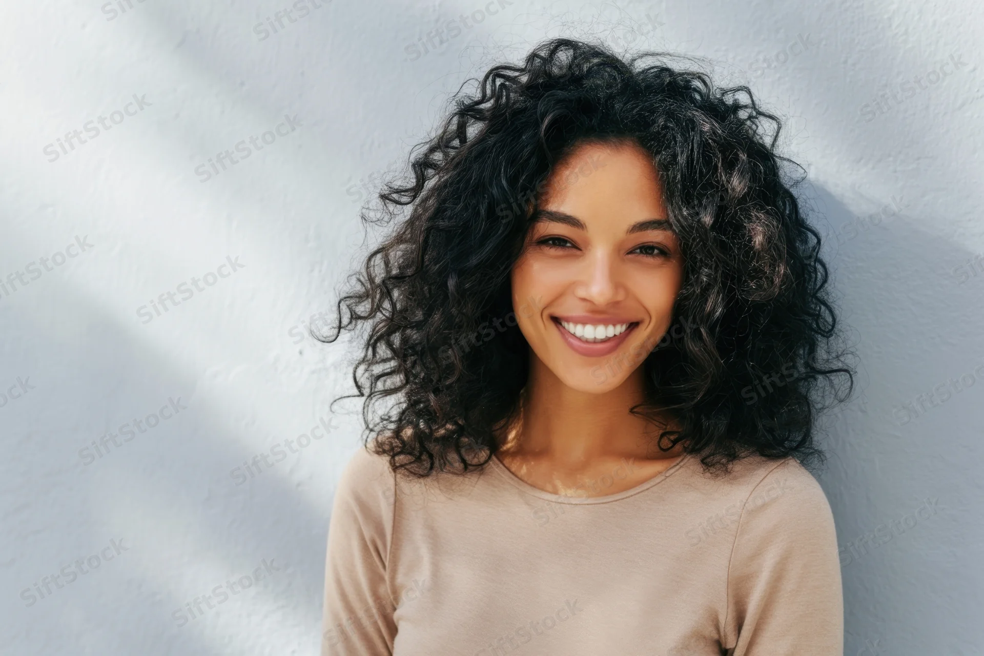 Smiling woman with curly black hair wearing beige top against white wall in sunlight