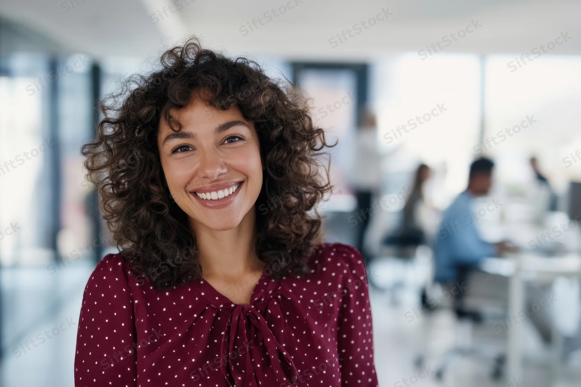 Smiling woman with curly hair in burgundy polka dot blouse standing in modern office environment