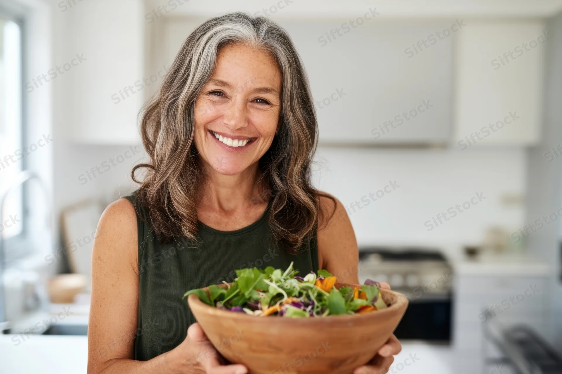 Smiling senior woman holding a wooden bowl of salad in a bright kitchen