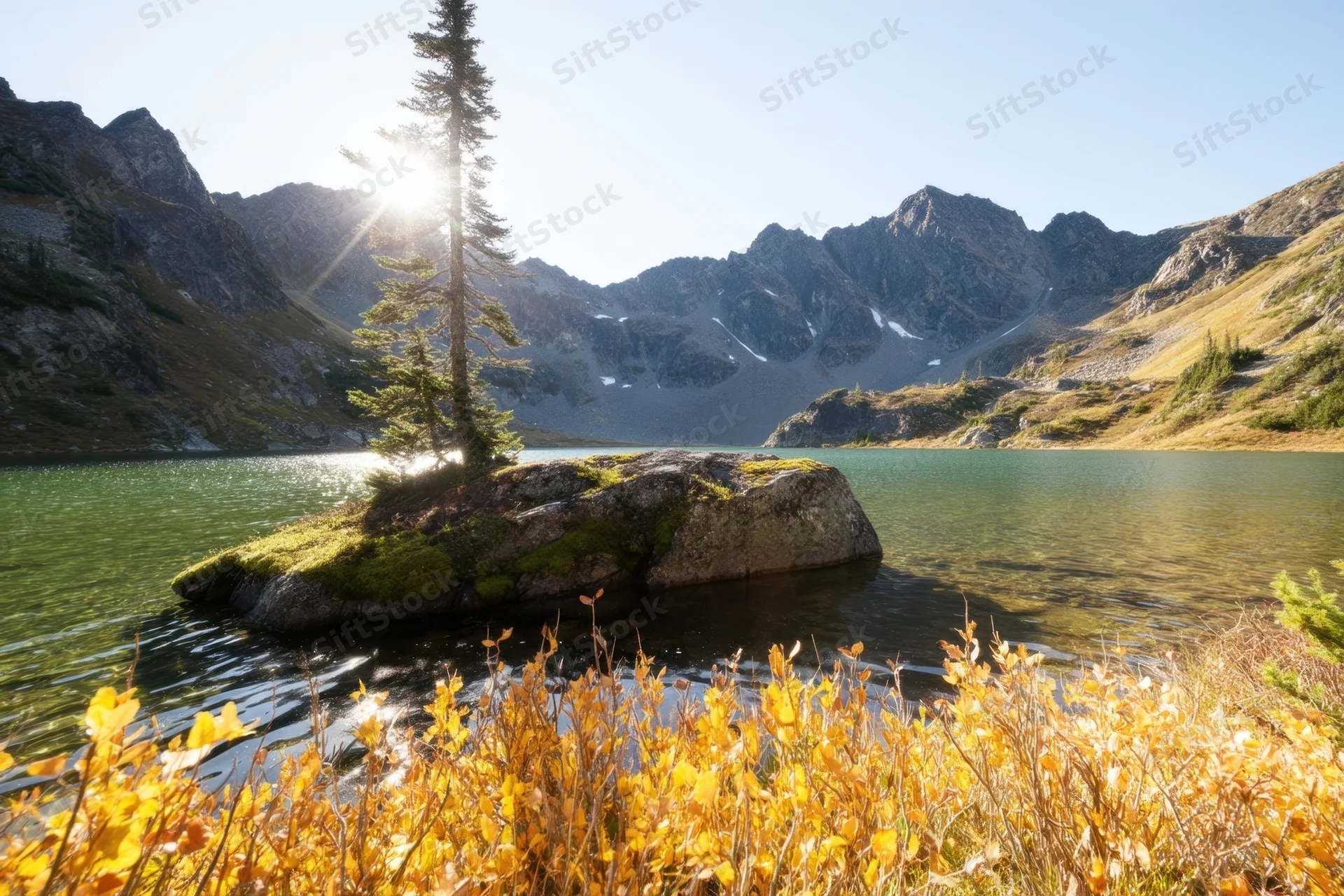 Sunlit alpine lake with golden autumn foliage in foreground and rocky mountains in background