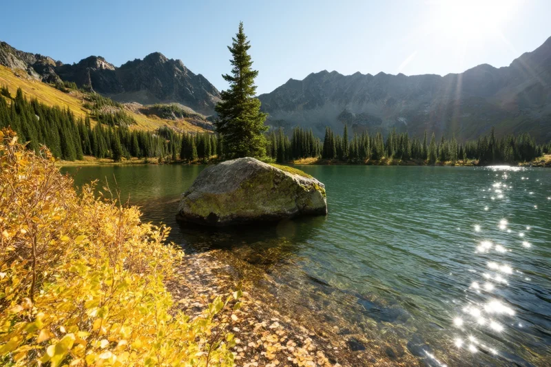 Sunlit alpine lake with golden autumn foliage and a rock in the foreground, surrounded by pine trees and mountains