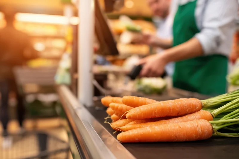 Fresh carrots on checkout counter with cashier in green apron scanning produce