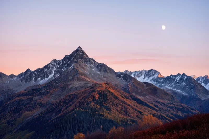 Alpine mountain range at sunset with snow-capped peaks and forested slopes under a twilight sky