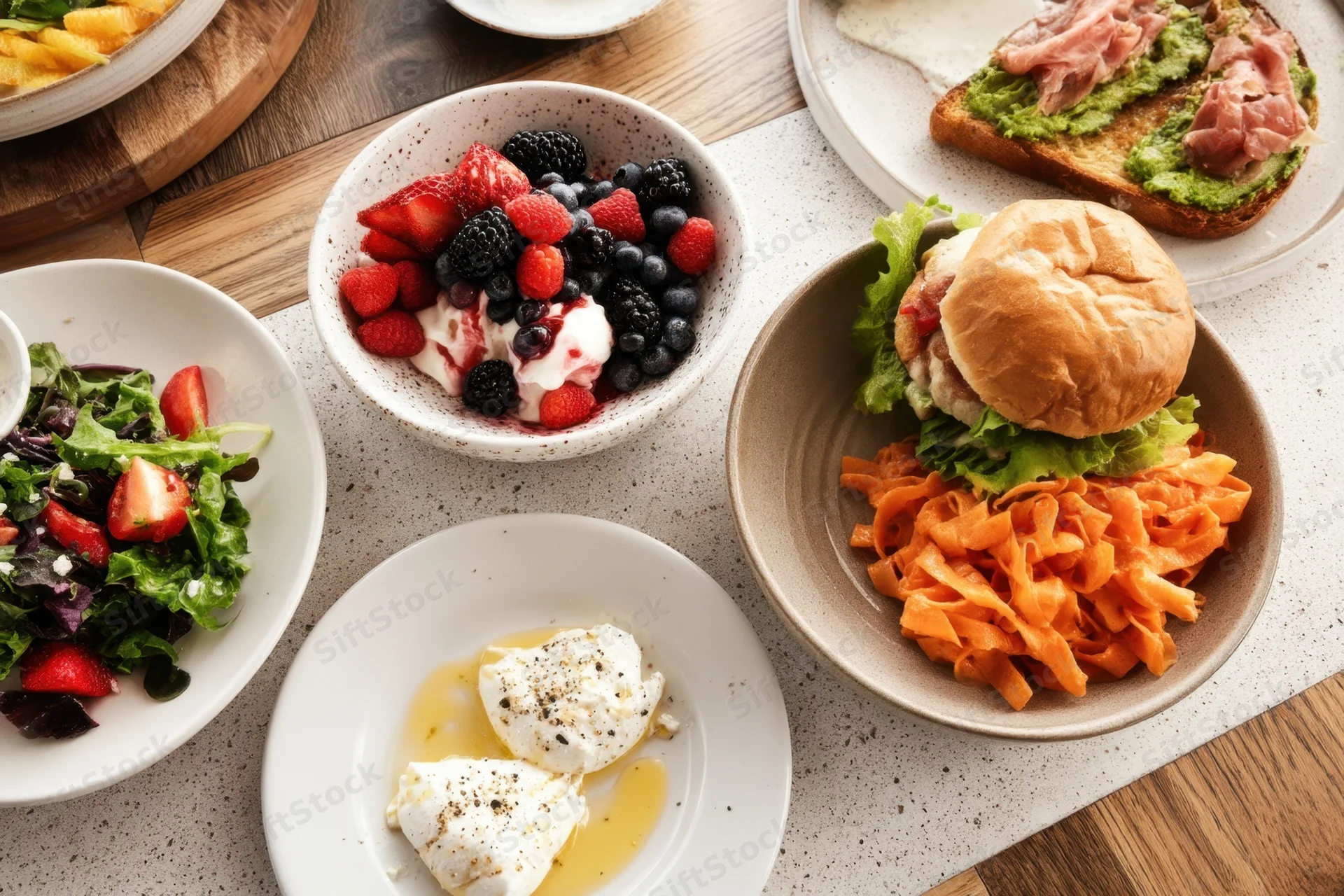Top-down view of breakfast dishes including avocado toast, burger, salad, berries and poached eggs on a kitchen countertop