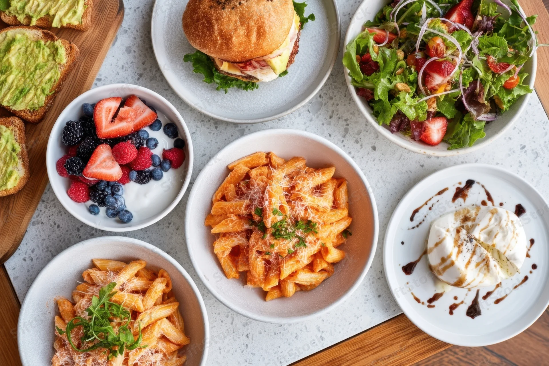 Top-down view of assorted dishes including pasta, salad, avocado toast, berries, and dessert arranged on a table