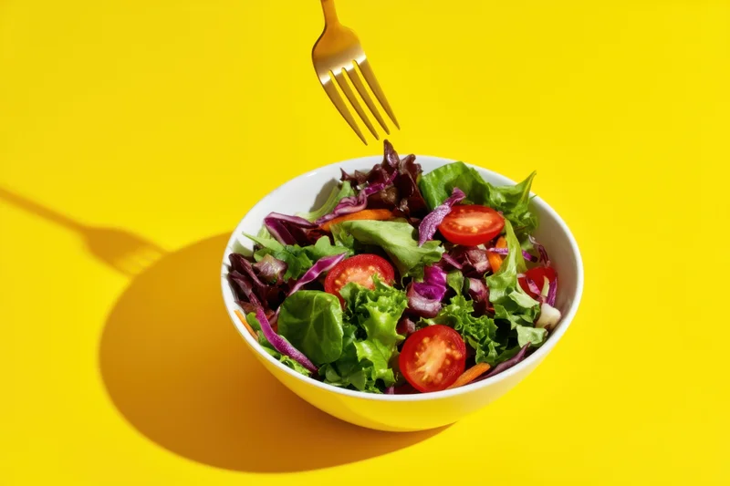 A white bowl of mixed salad with greens and cherry tomatoes on a yellow surface, a fork hovering above it