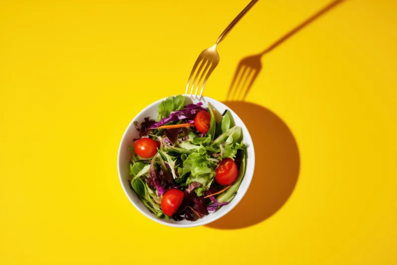 A white bowl of mixed salad with greens and cherry tomatoes on a yellow background, viewed from above with a fork inserted and casting a shadow