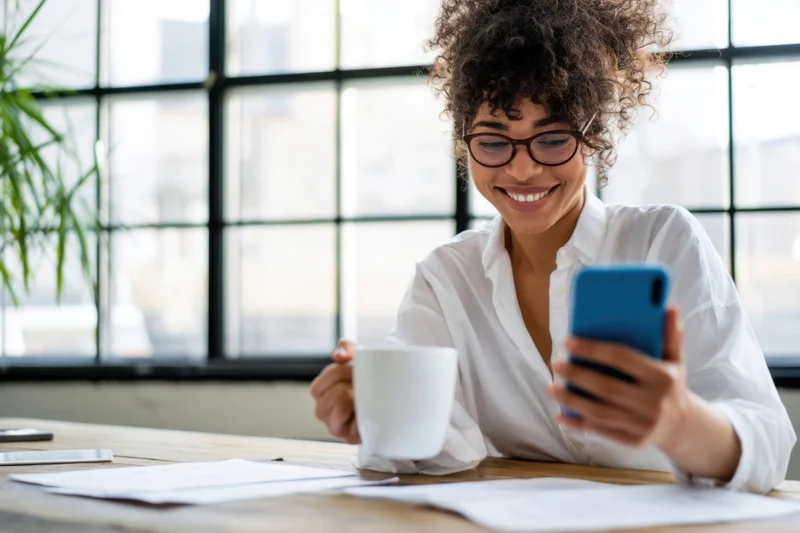 Woman smiling while using smartphone and holding coffee mug at wooden desk by window