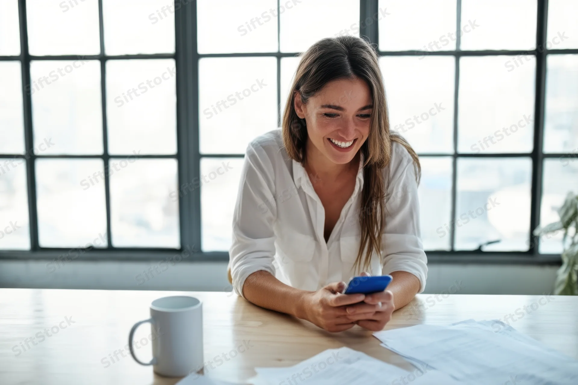 Woman smiling while using smartphone at wooden desk in bright office with large windows