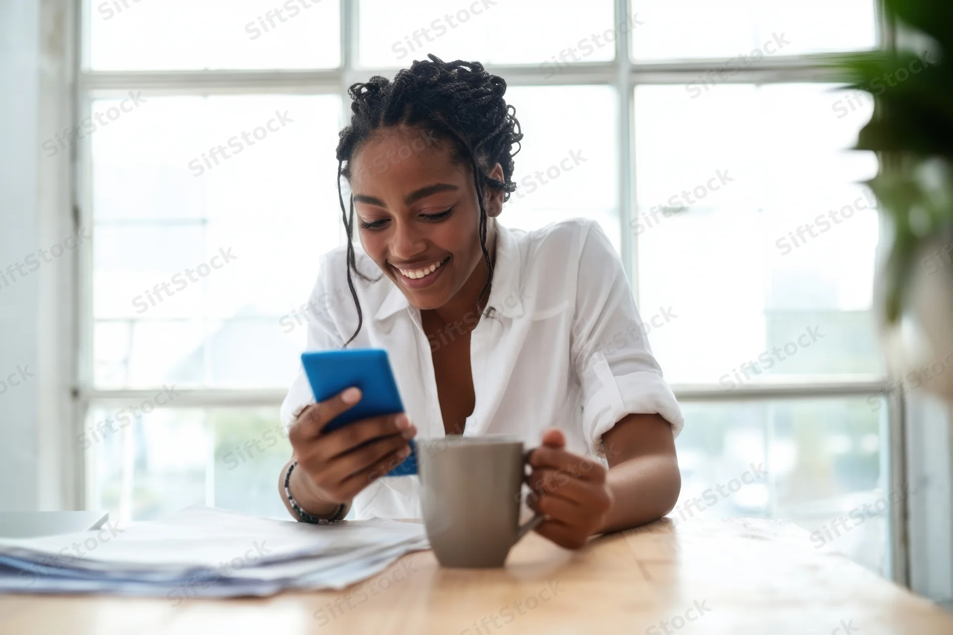 Woman smiling while using smartphone and holding coffee mug at wooden desk by window