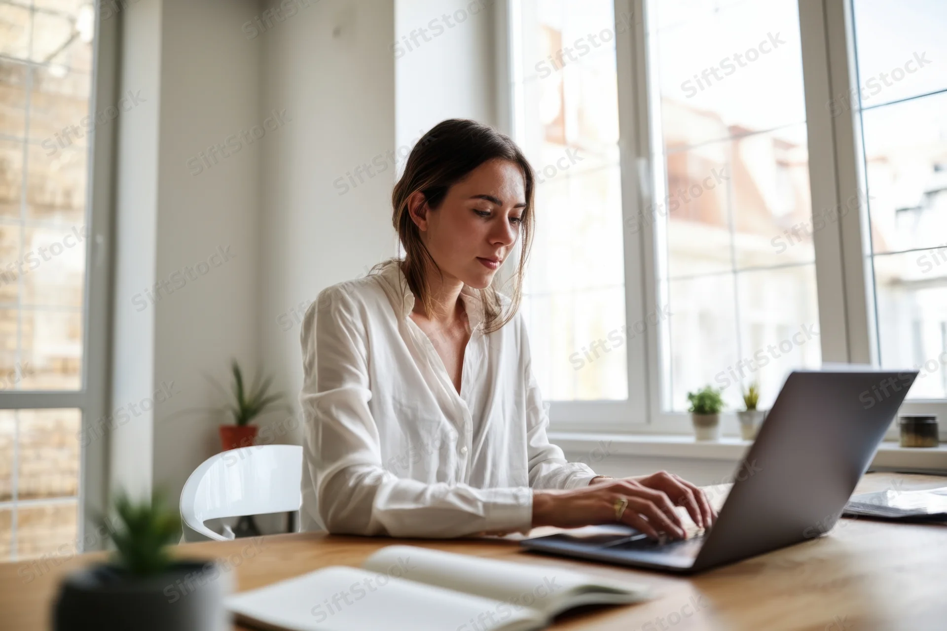Woman typing on laptop at wooden desk by window in bright office