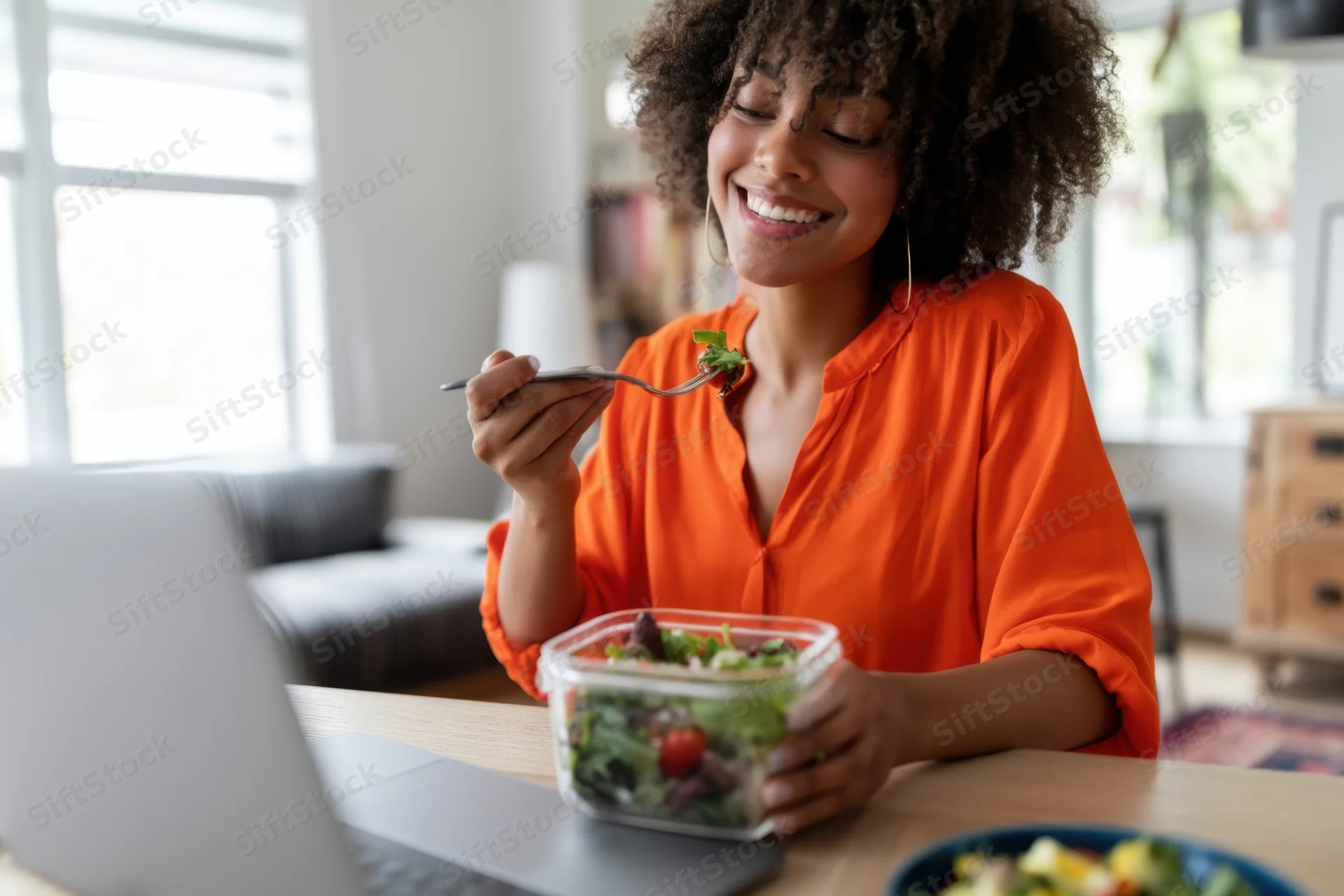 Woman eating salad from container at desk with laptop smiling