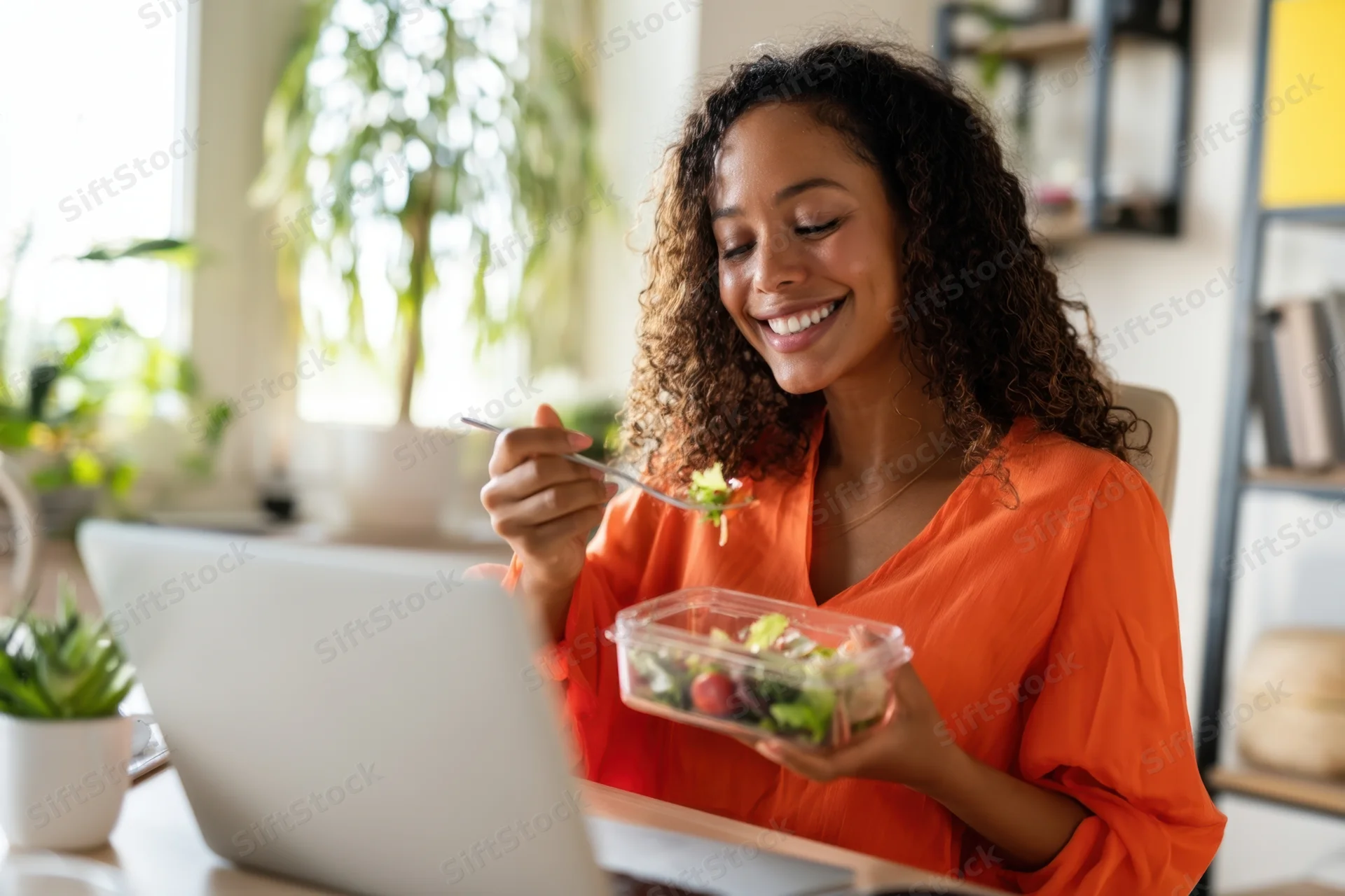 Woman eating salad from container while working on laptop at desk