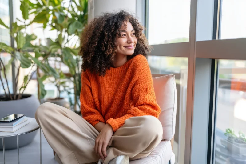 Woman with curly hair sitting cross-legged on cushioned chair by window smiling indoors