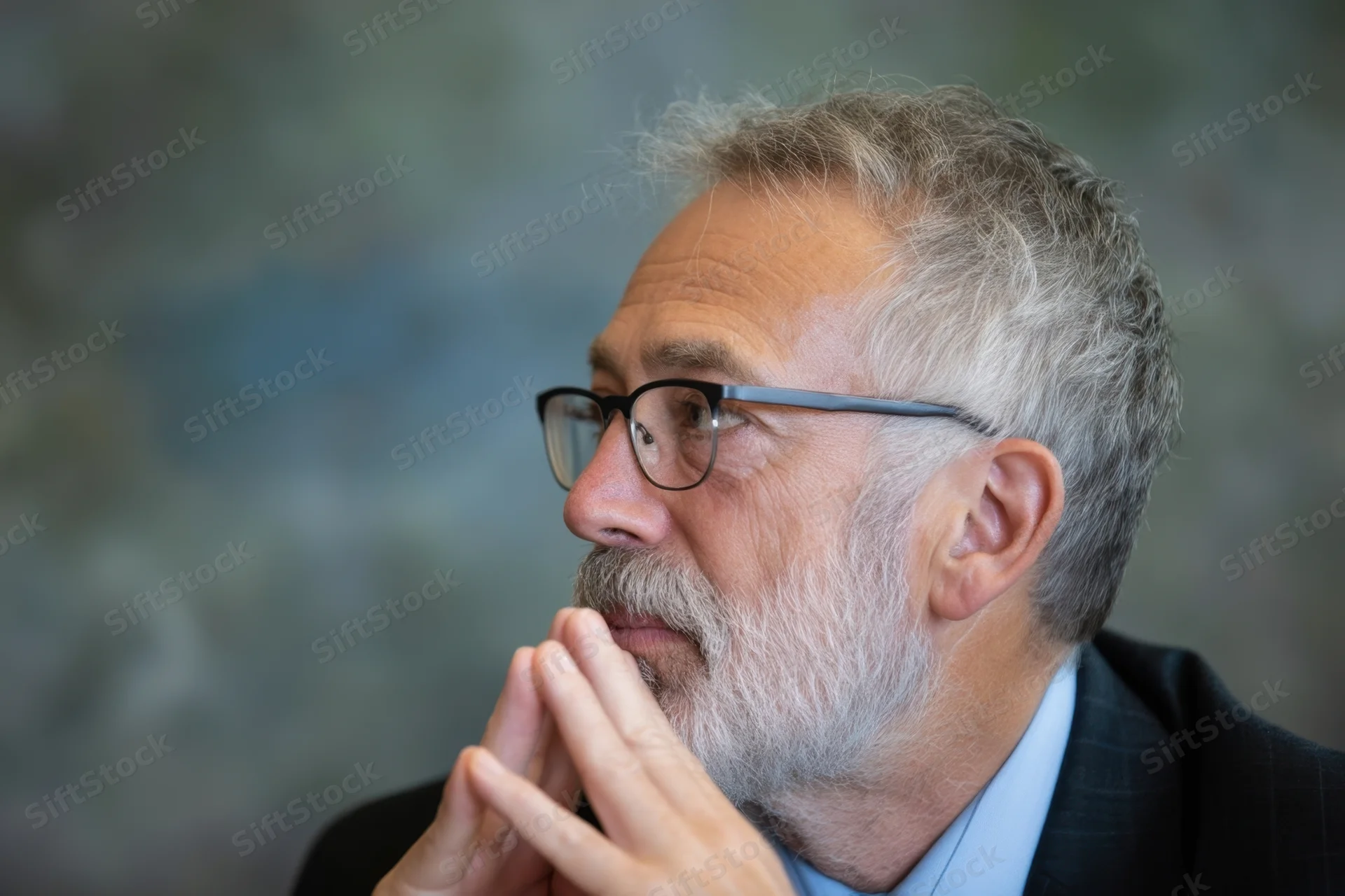 Senior man in profile with gray beard and glasses, hands clasped near chin, wearing suit and blue shirt in office setting