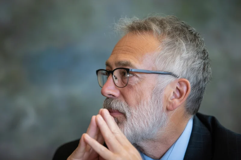 Senior man in profile with gray beard and glasses, hands clasped near chin, wearing suit and blue shirt in office setting