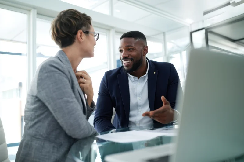 Two colleagues smiling and talking at a desk in a modern office