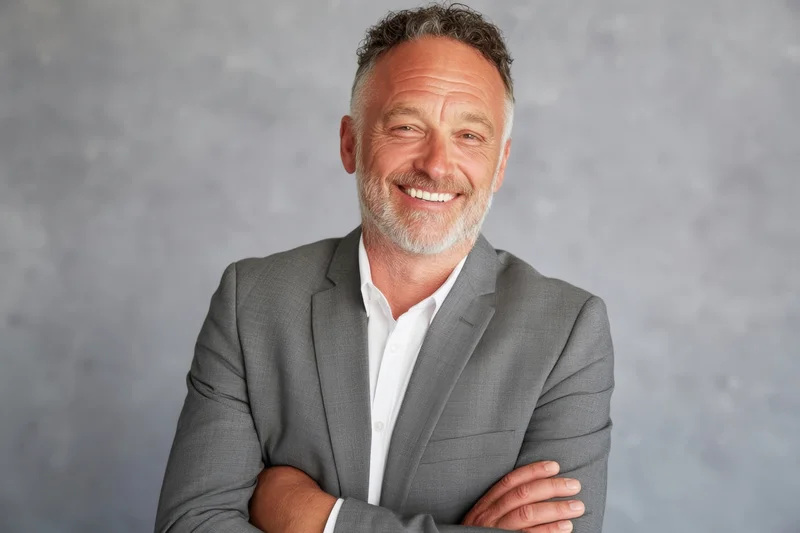 Middle-aged man in gray suit with white shirt smiling with arms crossed against gray background