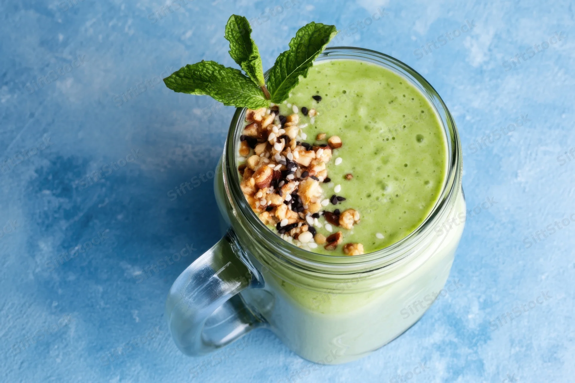 Pale green smoothie in a glass mason jar topped with nuts, seeds and mint on a blue surface