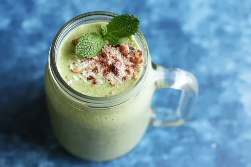 Pale green smoothie in a glass mason jar topped with mint and crumble on a blue surface