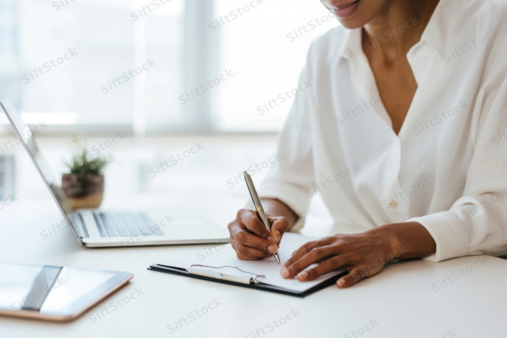 Woman writing on clipboard at desk with laptop and tablet in office