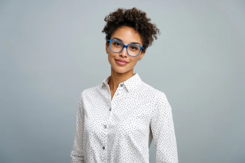 Woman with short curly hair and blue-framed glasses wearing a white polka dot shirt in a studio setting