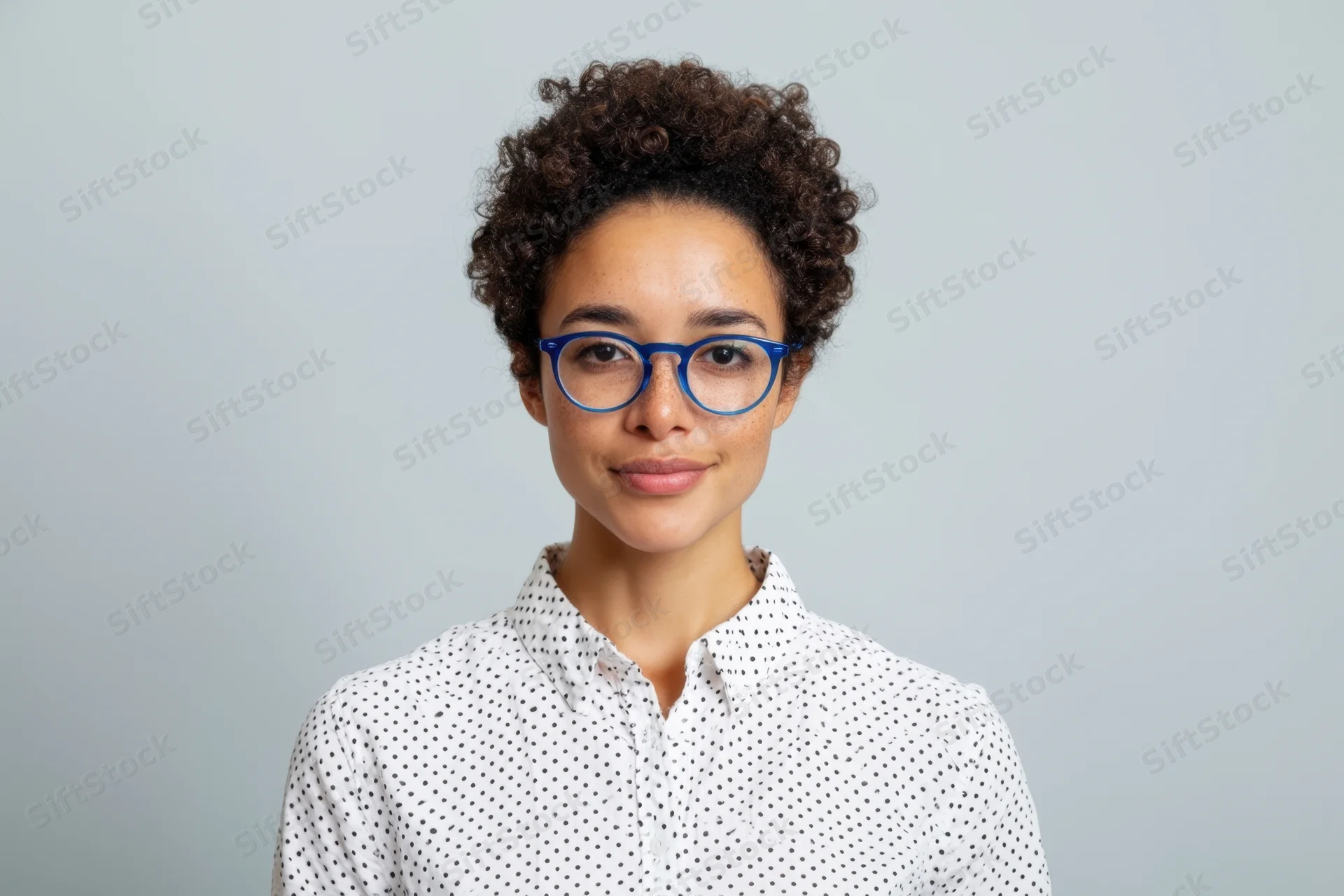 Woman with short curly hair and blue-framed glasses wearing a white polka dot shirt in a studio portrait