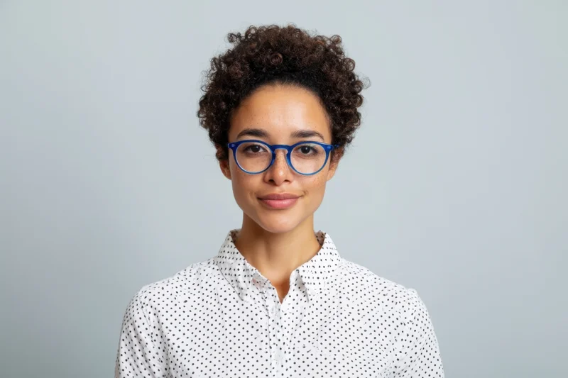 Woman with short curly hair and blue-framed glasses wearing a white polka dot shirt in a studio portrait