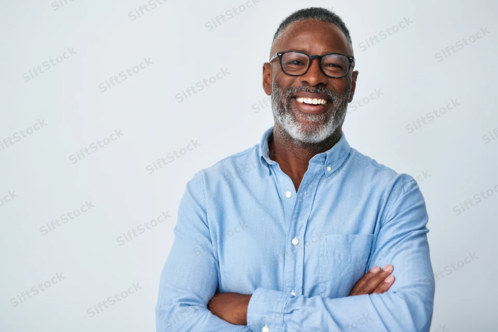 Smiling man with beard and glasses wearing blue shirt, arms crossed, in studio setting