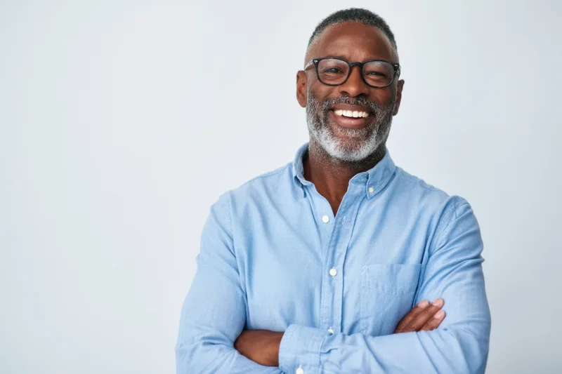 Smiling man with beard and glasses wearing blue shirt, arms crossed, in studio setting