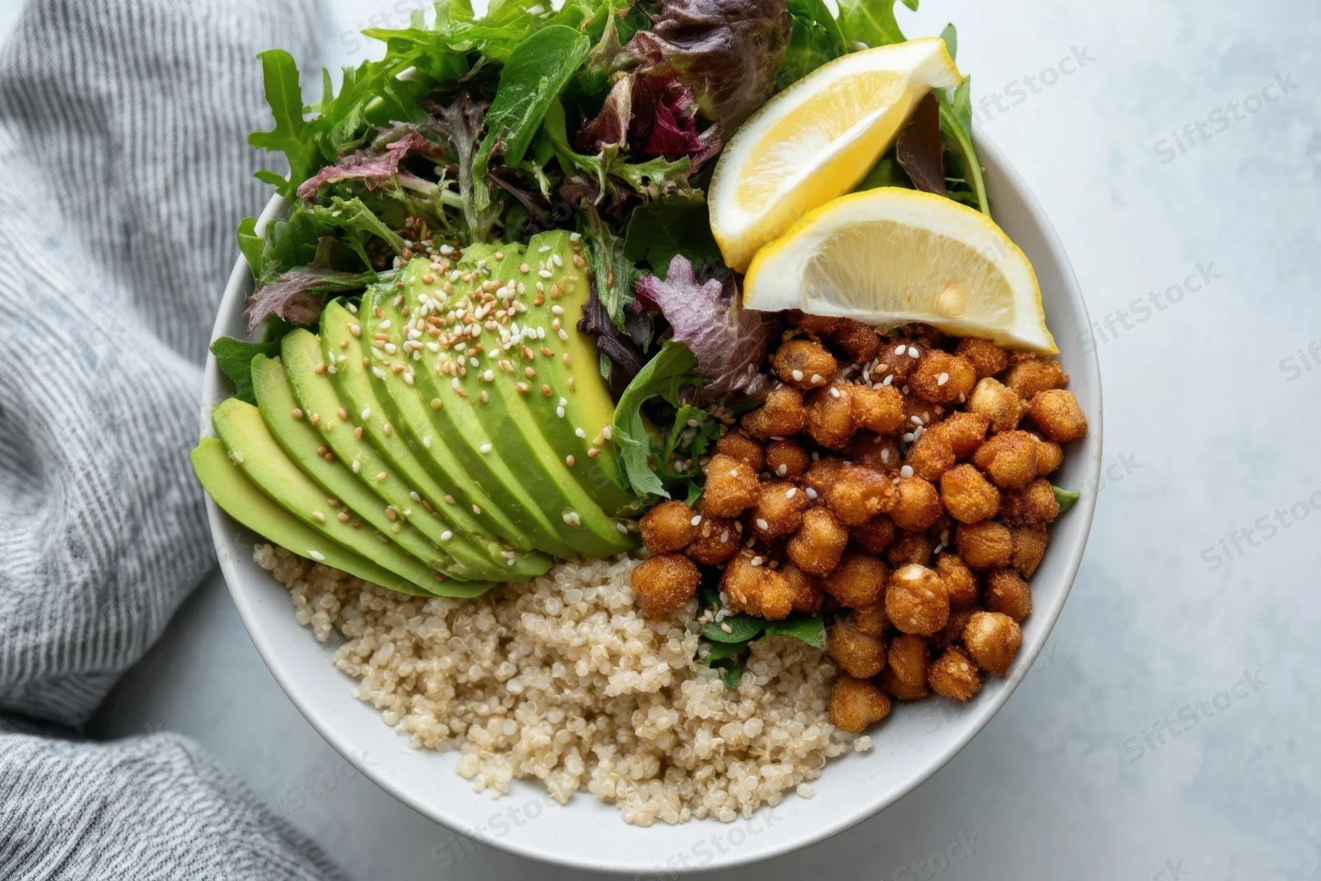 A salad bowl with sliced avocado, quinoa, crispy chickpeas, lemon wedges, and mixed greens on a white surface beside a gray cloth