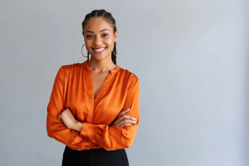 Confident woman with braided hair and hoop earrings smiling with arms crossed in orange blouse against light gray background