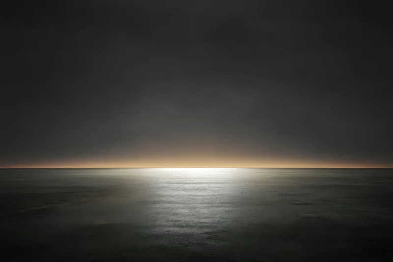 Calm ocean surface reflecting a glowing horizon at dusk under dark clouds