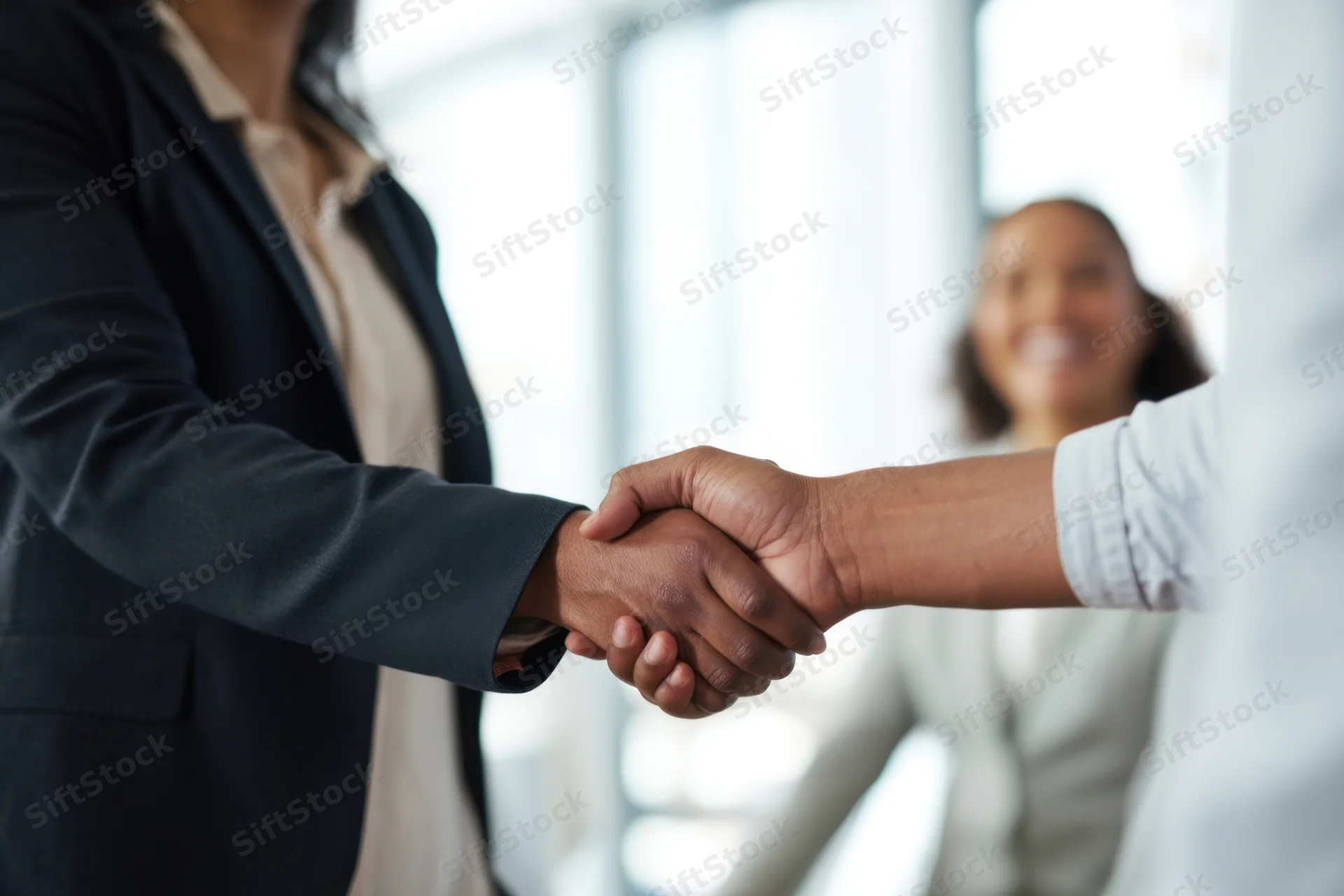 Two businesswomen shaking hands in an office setting with a smiling colleague in the background