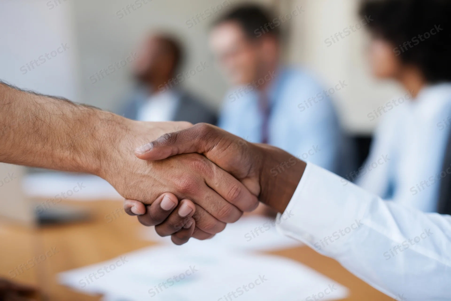 Closeup of two hands shaking over a desk at a business meeting with colleagues in the background