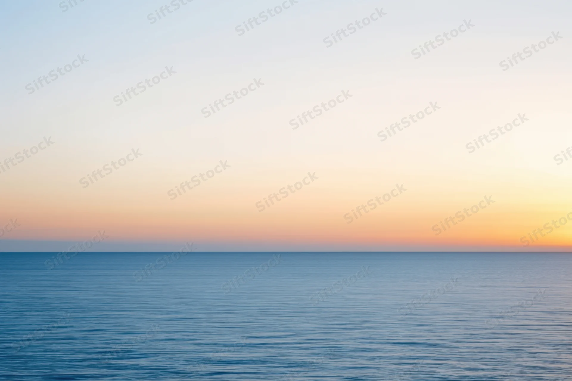 Calm ocean surface under a hazy sunset sky with peach and gold tones