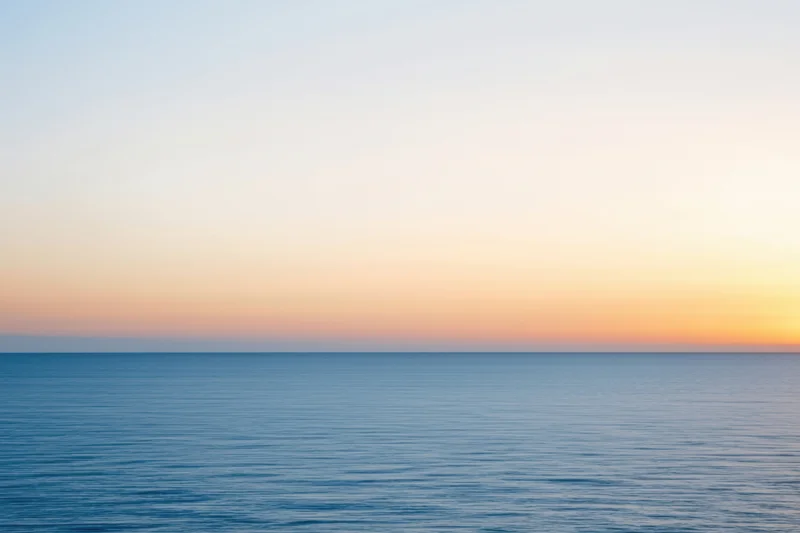 Calm ocean surface under a hazy sunset sky with peach and gold tones
