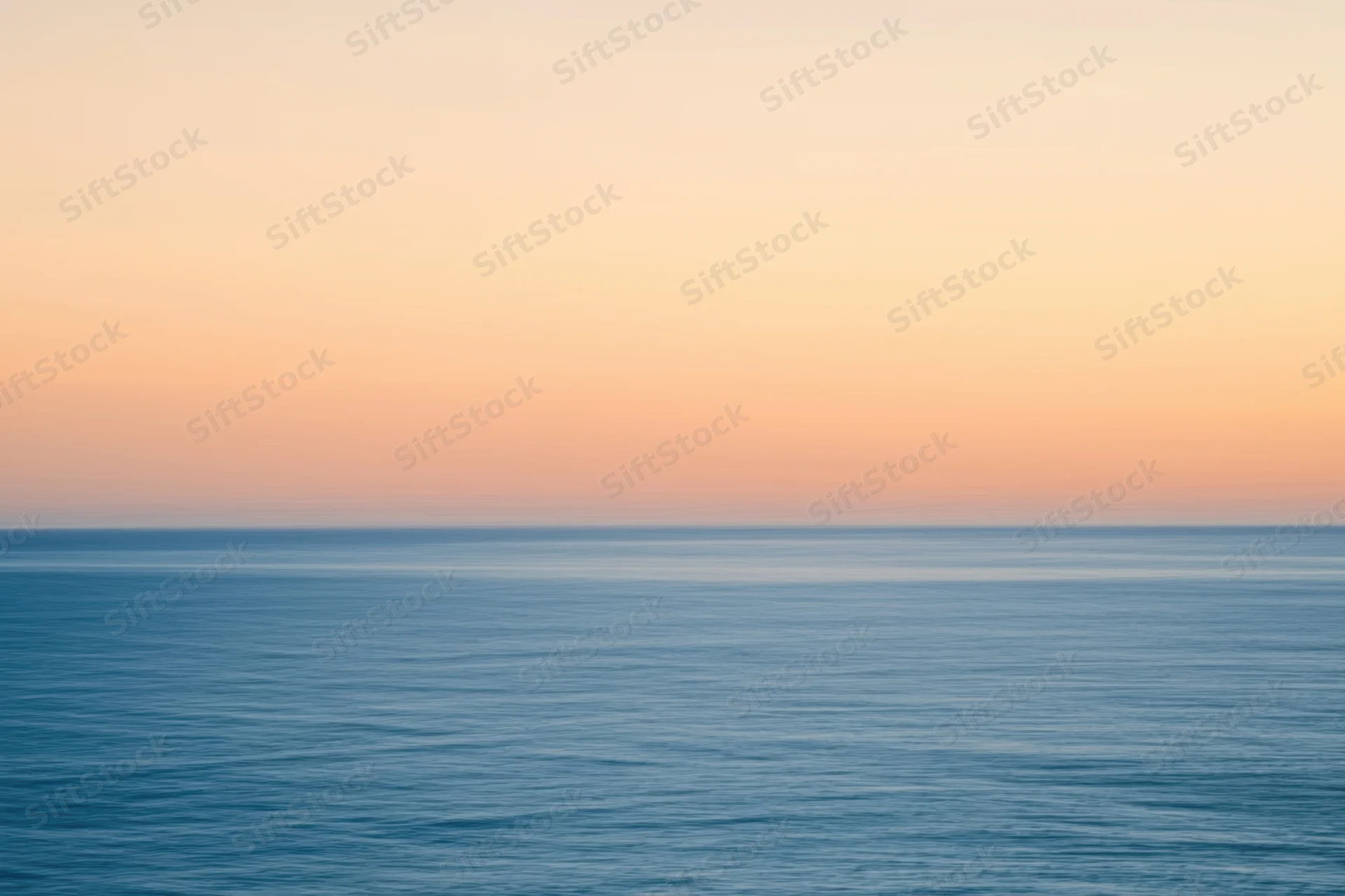Calm ocean surface under a hazy peach and gold sunset sky