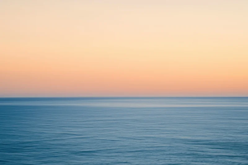 Calm ocean surface under a hazy peach and gold sunset sky