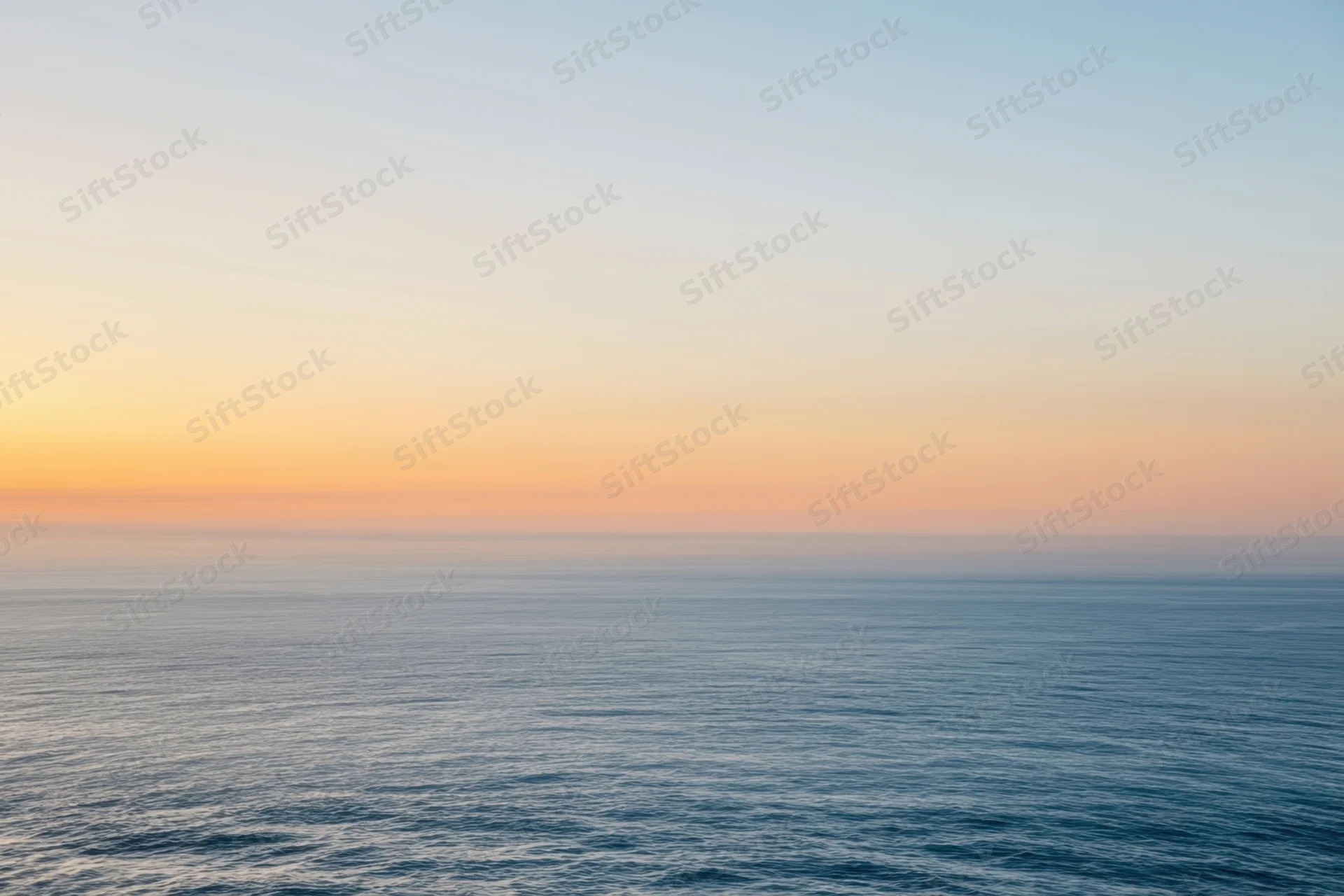 Calm ocean surface under a hazy sunset sky with peach and gold tones