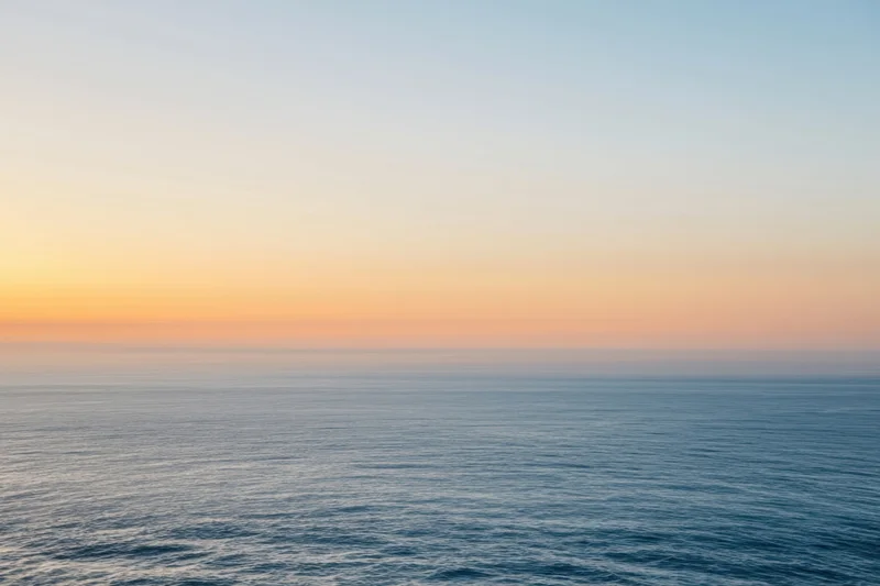 Calm ocean surface under a hazy sunset sky with peach and gold tones