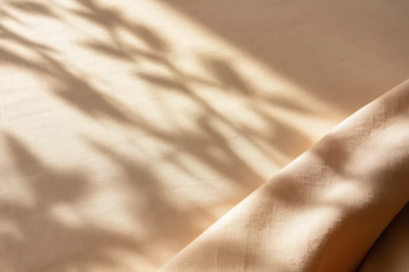 Beige fabric with dappled leaf shadows cast by sunlight, folded and textured, in a calm indoor setting