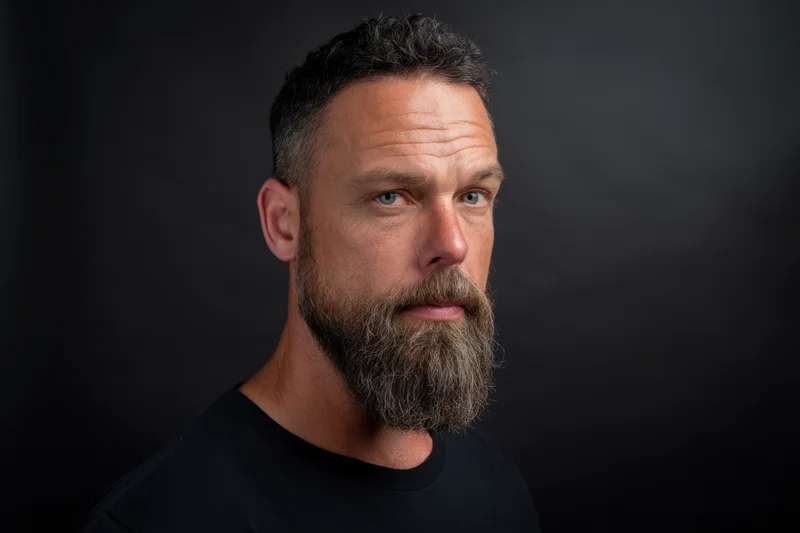 Man with full beard and short dark hair in black shirt against dark background, looking directly at camera with serious expression
