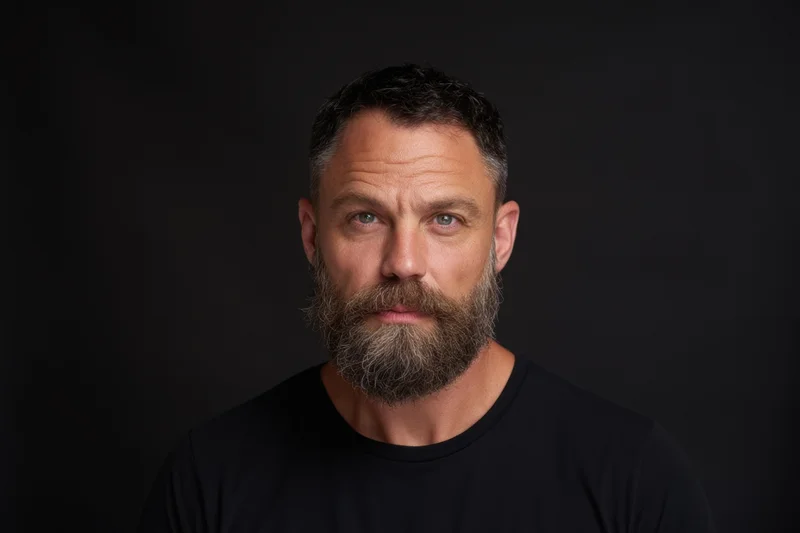 Man with full beard and short dark hair in black tshirt looking directly at camera against dark background