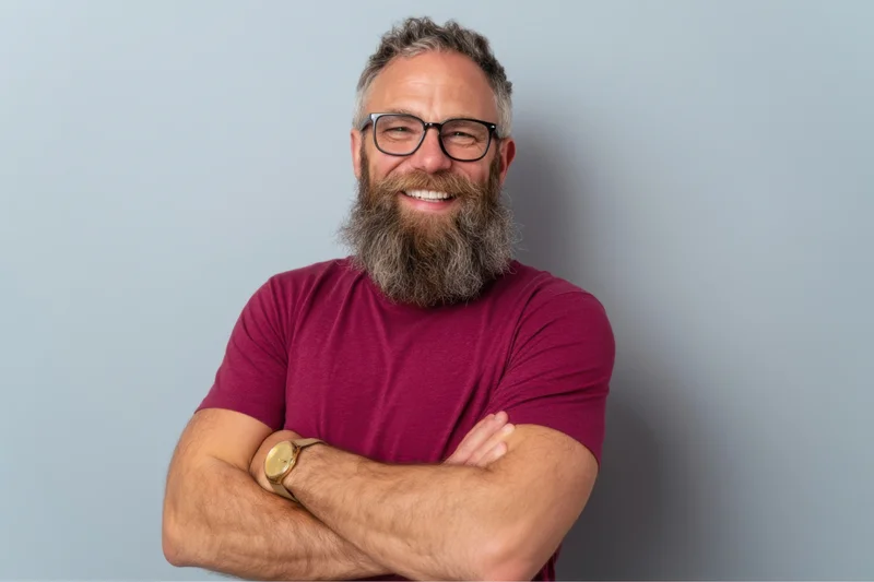 Middle-aged man with full beard and glasses smiling with arms crossed against light blue background