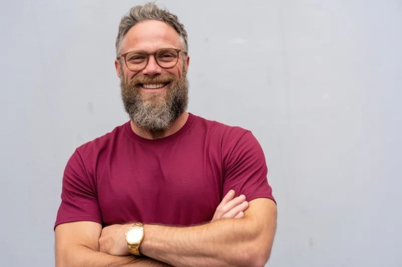 Middle-aged man with full beard and glasses smiling with arms crossed against light gray background