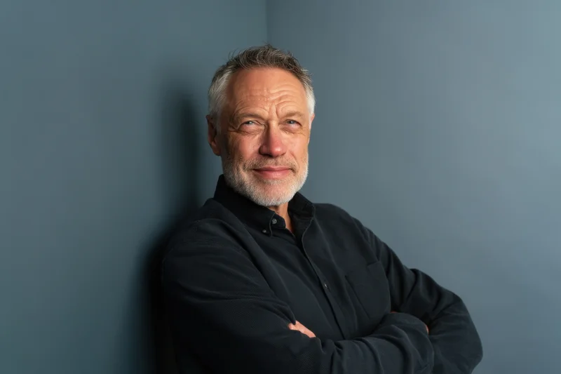 Middle-aged man with salt-and-pepper hair and beard leaning against a gray wall with arms crossed, wearing a black shirt, looking directly at camera