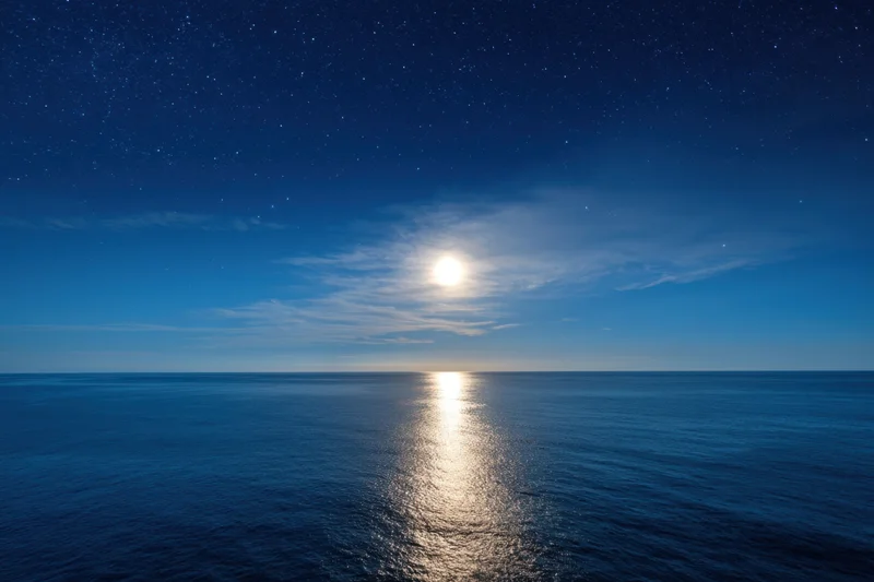 Full moon glowing over calm ocean at night with stars in sky and reflection on water