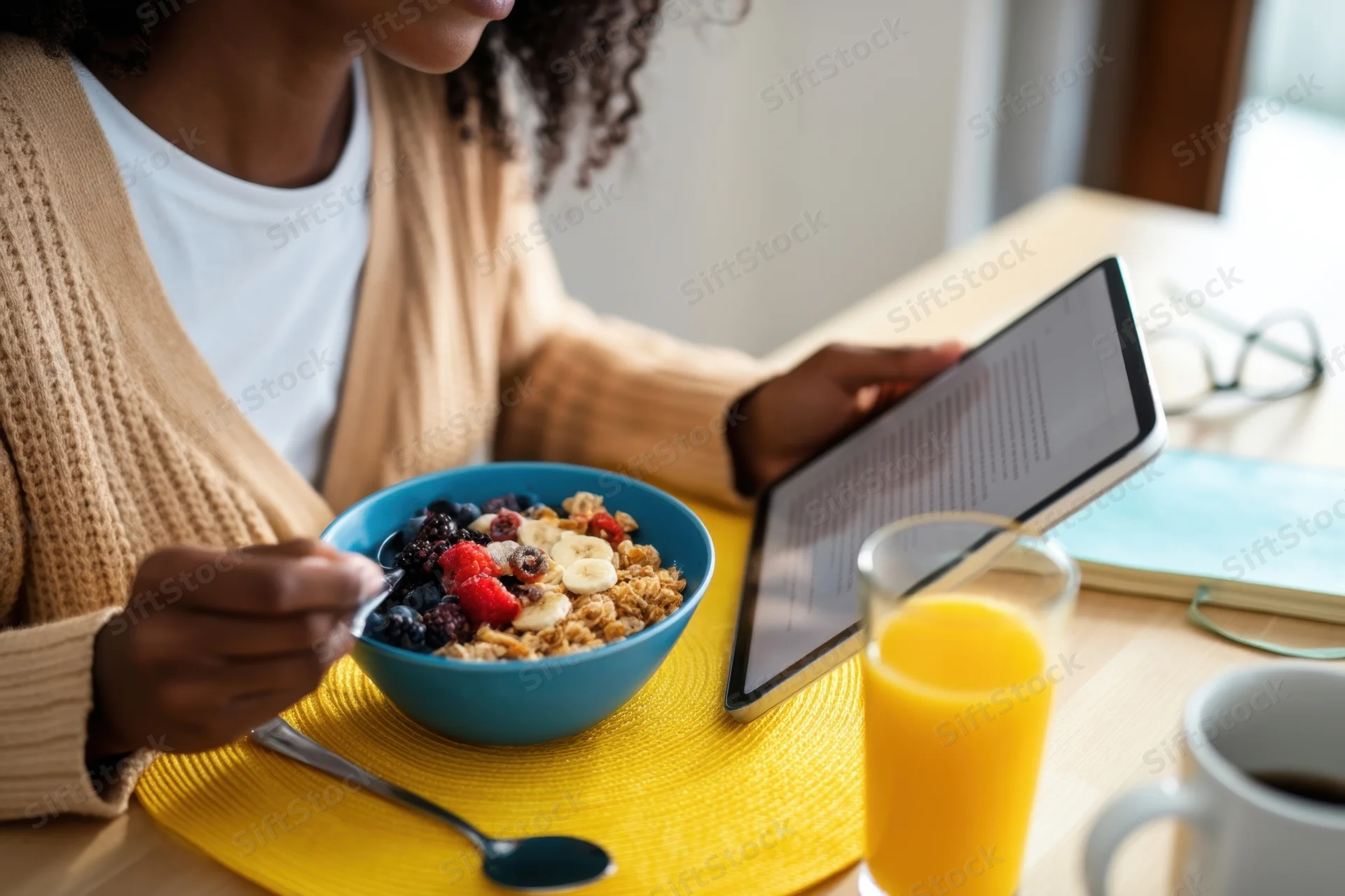 Woman eating breakfast with fruit and granola while reading on a tablet at a wooden table